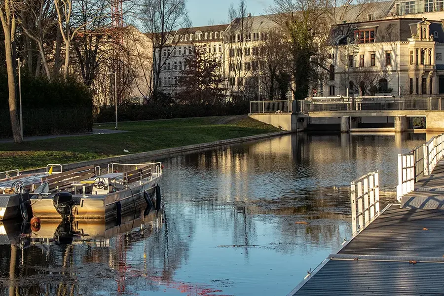 Zwei leere Motorboote liegen an einem Steg am stadthafen Leipzigauf einem ruhigen Kanal, umgeben von Bäumen und historischen Gebäuden im Hintergrund. Ein Baukran ragt über die Szenerie hinaus.