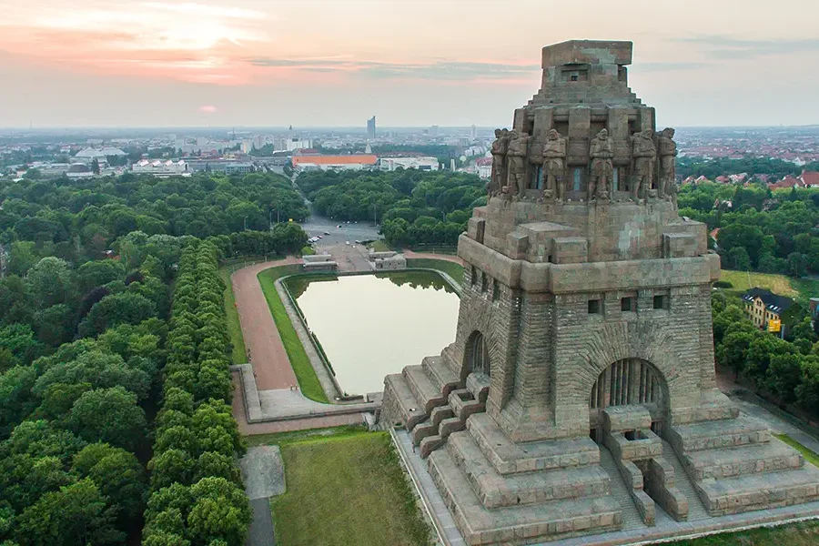 Luftaufnahme des Völkerschlachtdenkmals in Leipzig, umgeben von Bäumen und einer Wasserfläche, im Hintergrund die Stadtlandschaft bei Sonnenuntergang.