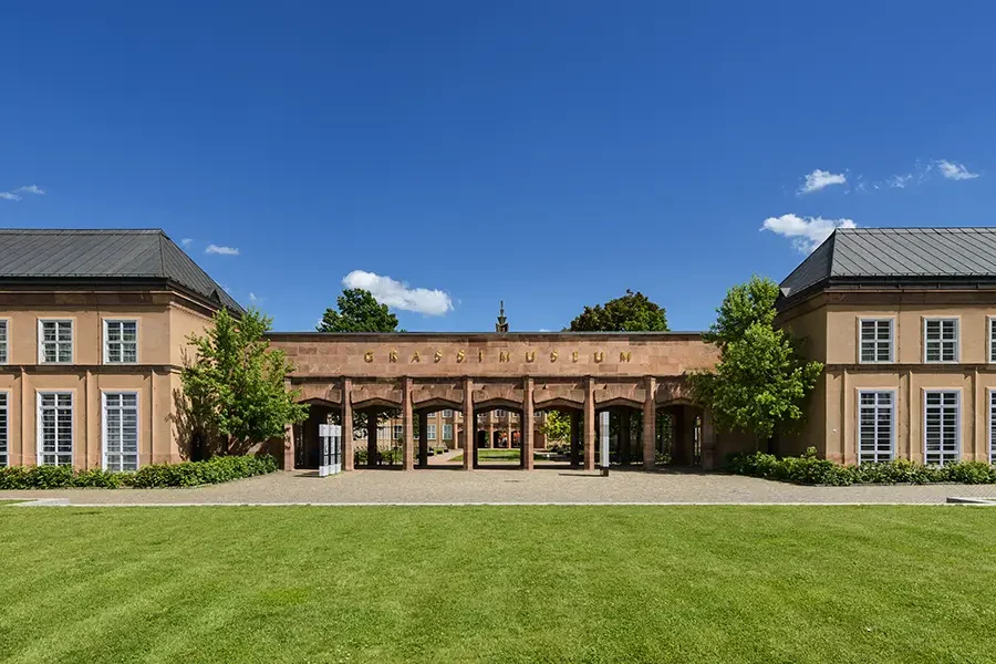 Außenansicht des Grassimuseums in Leipzig, mit einem zentralen Eingangsbereich und zwei symmetrischen Gebäudeflügeln, umgeben von grünen Rasenflächen und Bäumen unter klarem, blauem Himmel.