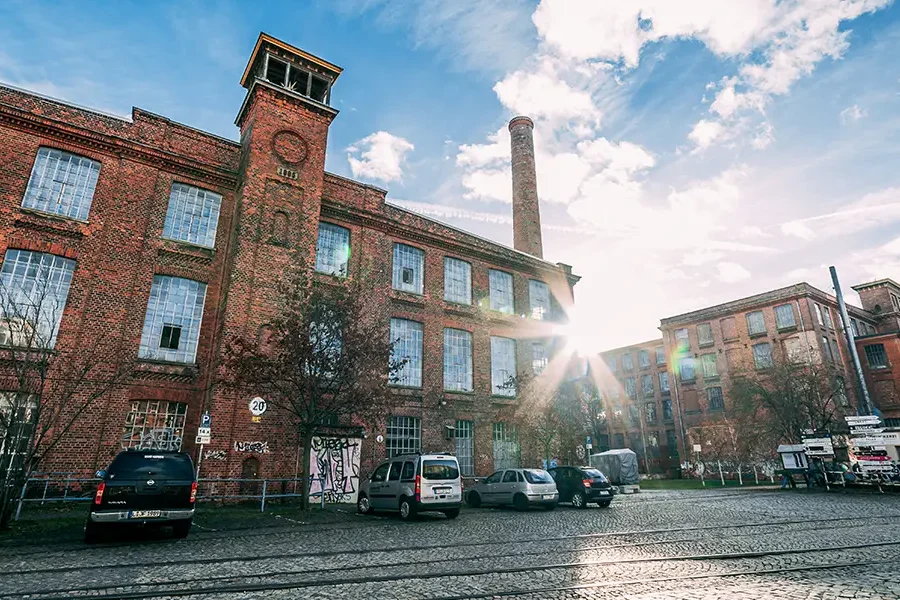 Historisches Backsteingebäude der Leipziger Baumwollspinnerei mit hohen Fenstern, einem Turm und einem Schornstein, beleuchtet von Sonnenstrahlen unter einem blauen Himmel.